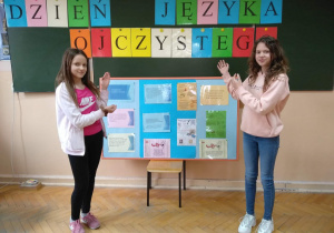 Dwie uczennicy przy tablicy prezentują kolorowy napis i tablicę informacyjną dotyczącą obchodów Dnia Języka Ojczystego